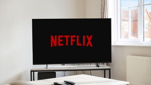Una habitación con una televisión mostrando 'NETFLIX', una mesa con controles remotos y una ventana con vista a edificios.