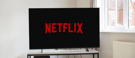 A room with a television displaying 'NETFLIX' on the screen, a table with remotes, and a window with a view of buildings.