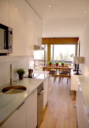 A modern kitchen with white cabinets, a stainless steel sink, and a dining area with wooden chairs and a table. - Beautiful apt. in Anzère in the Swiss alps. Amazing views across the mountains (Ayent)
