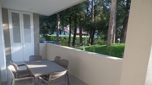 A balcony with a table and chairs, a white door with shutters, and a view of a green landscape with trees. - Well-equipped Two-room Flat in a new Residence (San Michele al Tagliamento)
