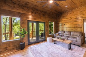 A wooden cabin interior with a sofa, a coffee table, a potted plant, and large windows. - Smoky Sunset Getaway -Brand New Beautiful Mountain Cabin (Sevierville)