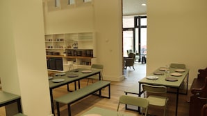 Un comedor con varias mesas y sillas, una zona de cocina con estantes llenos de vajilla y vistas al exterior a través de un ventanal grande.