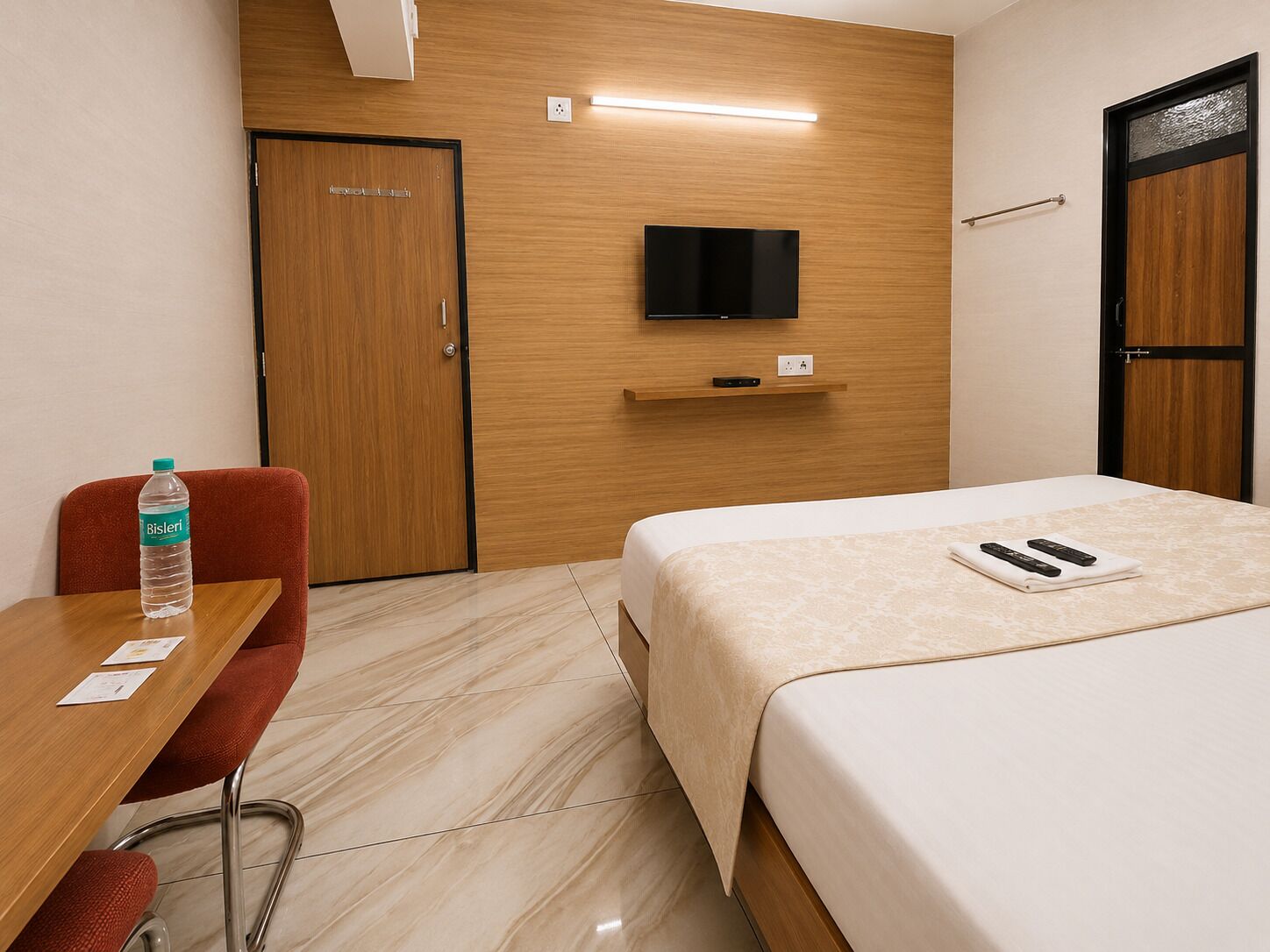A hotel room with a bed, a TV mounted on the wall, a desk with a chair, and a bottle of water on the desk.