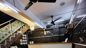 A modern interior with a marble floor, a reception desk, a staircase with glass railings, and ceiling fans. - Hotel Grand Riva (Rajkot)