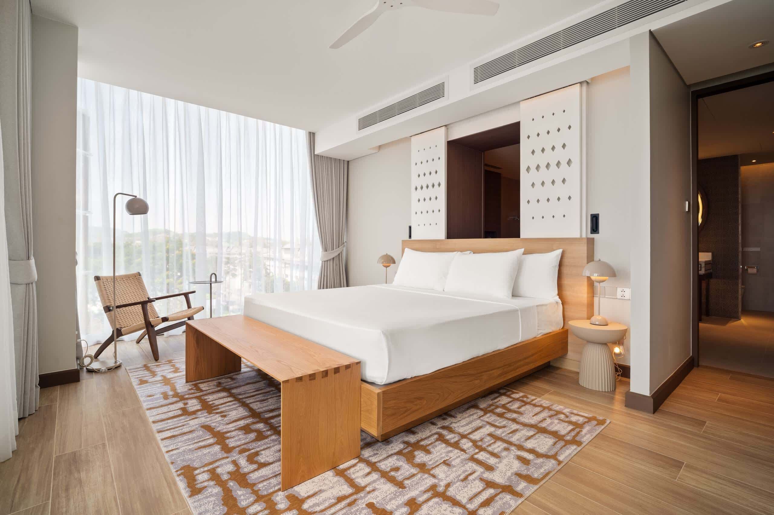 A modern hotel room with a large bed, a bedside table, a chair, and a view of the city.