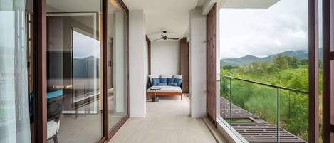 A balcony with a sofa and a table, a ceiling fan, and a view of greenery and mountains.