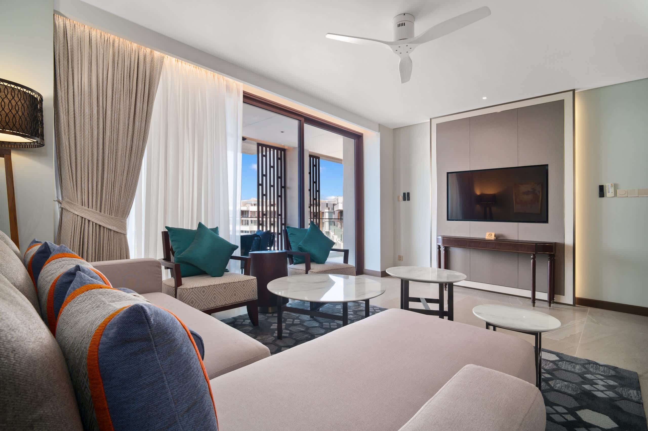 Quarto de hotel com sofá, duas poltronas, mesa de centro, televisão e ventoinha no teto.