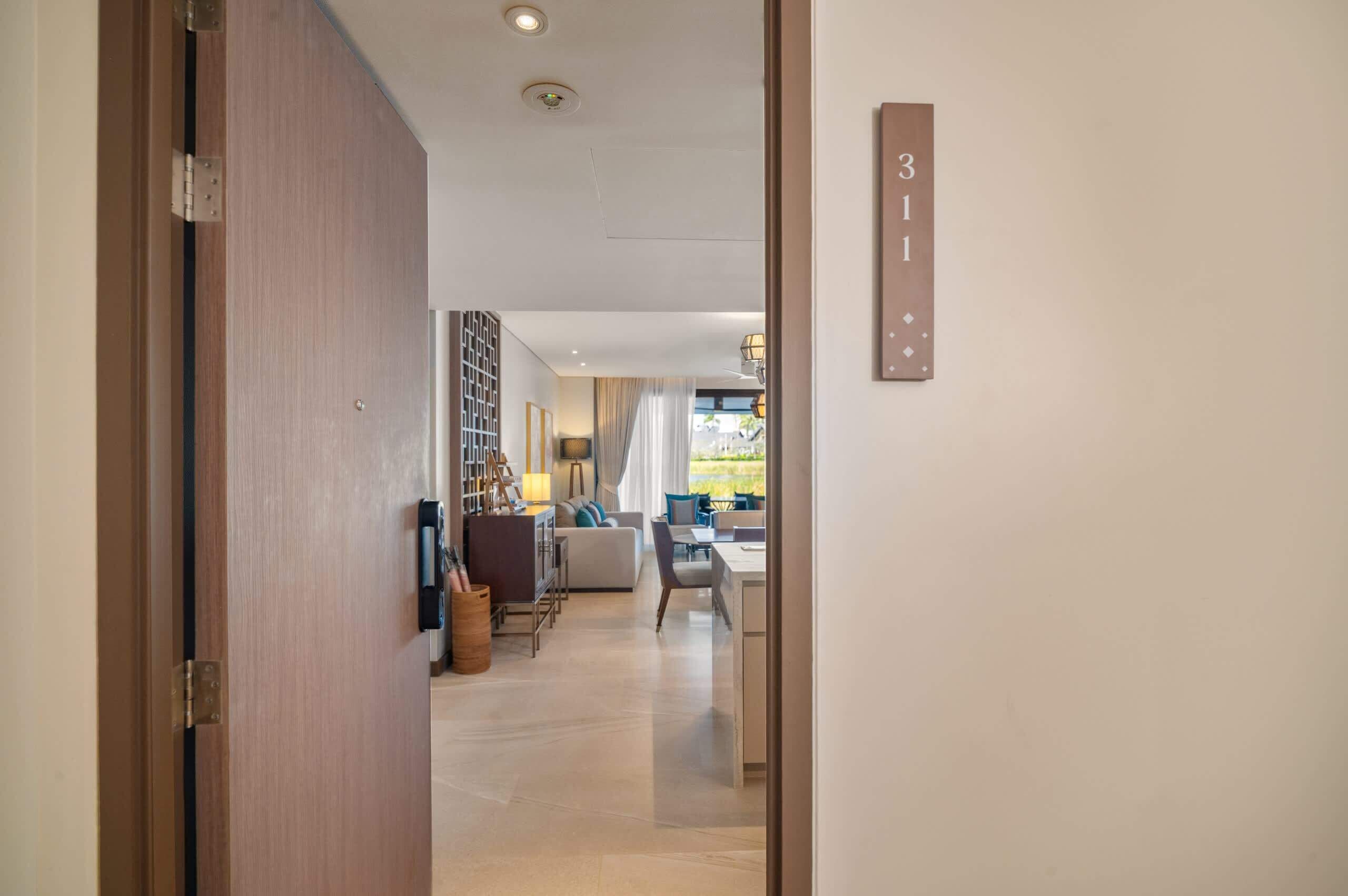 Un interior moderno con una puerta de madera, número de habitación visible, zona de comedor con mesa y sillas, y un sofá.