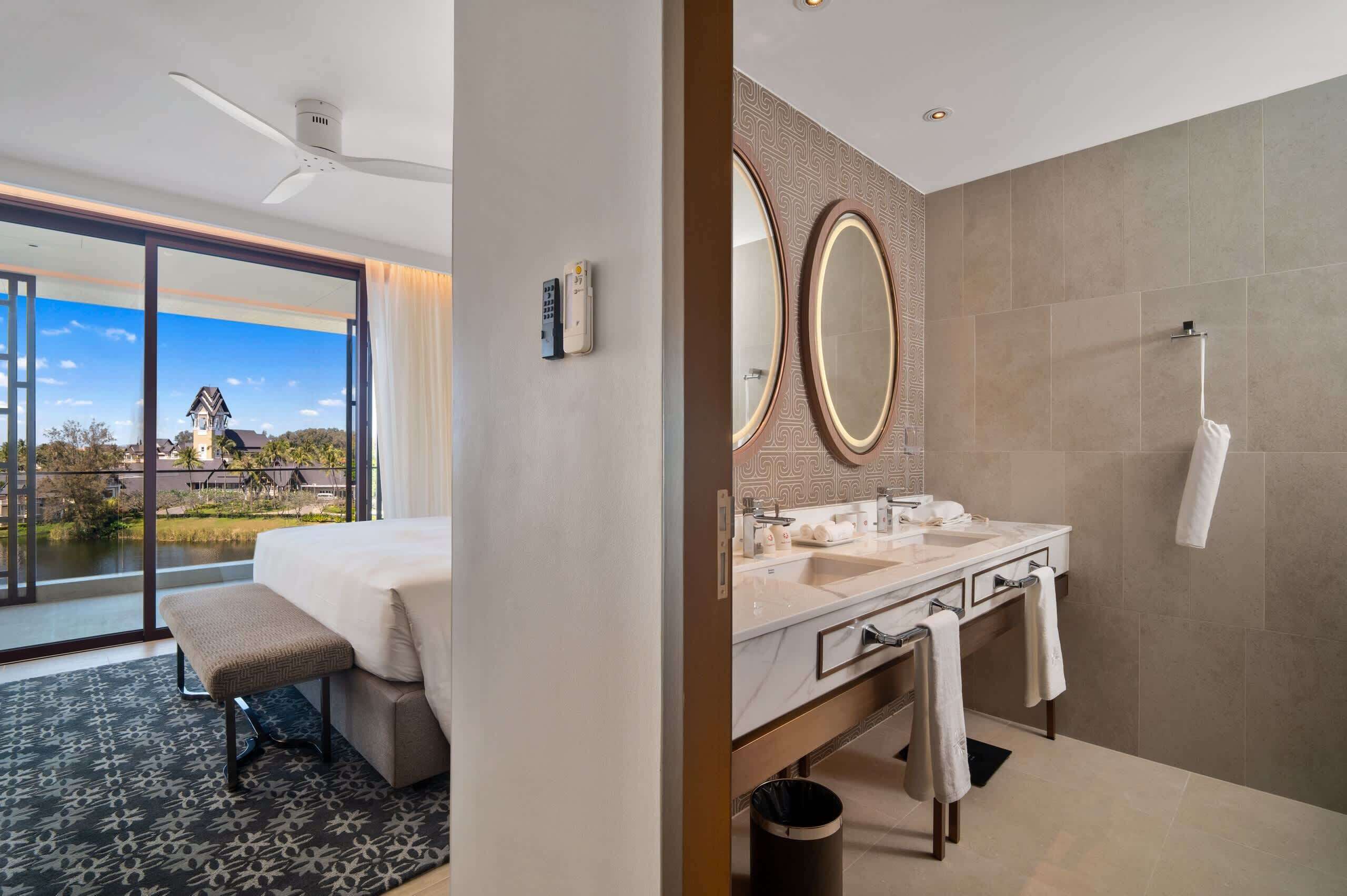 A hotel room with a large bed, a bench, a bathroom with a sink and mirror, and a view of a building and water outside.
