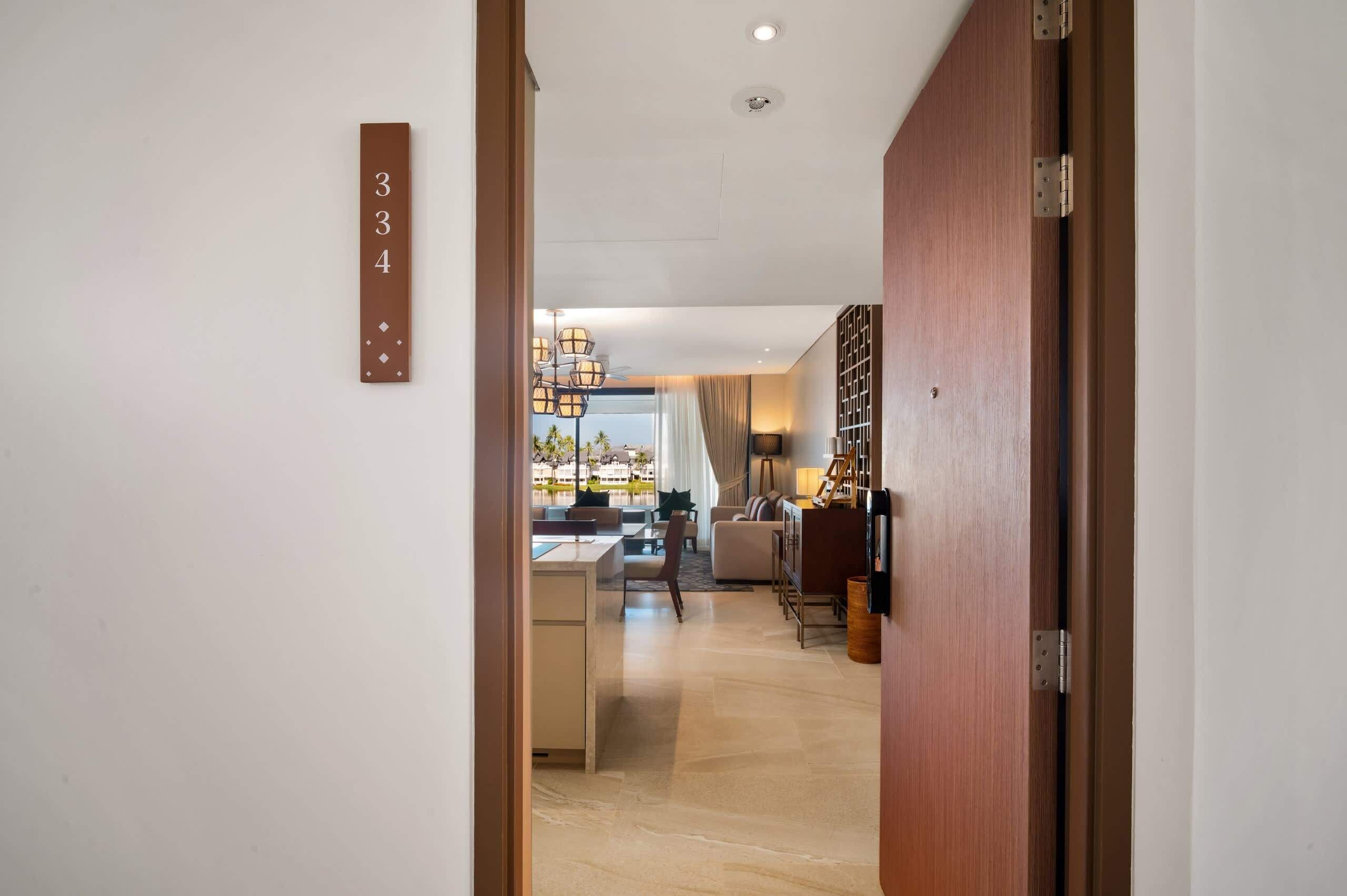 A modern hotel room with a wooden door, a visible number '334' on the wall, a dining area with a table and chairs, and a view of the outside.