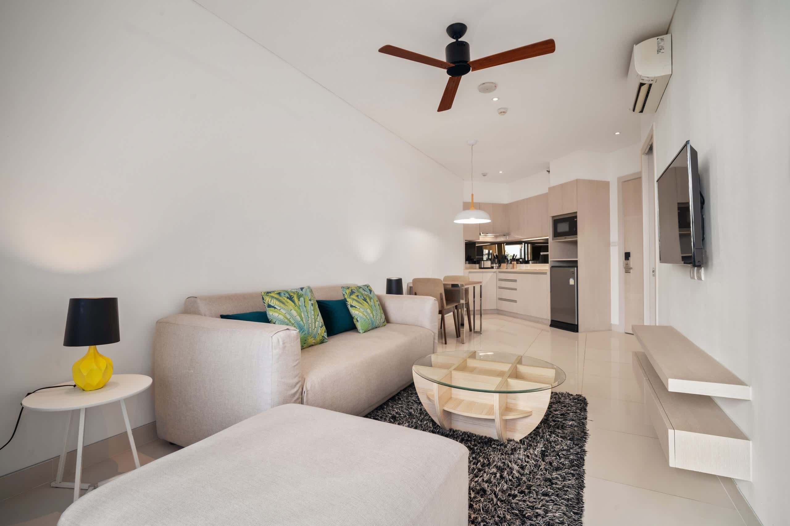 A modern living room with a sofa, a glass coffee table, a ceiling fan, and a kitchen in the background.