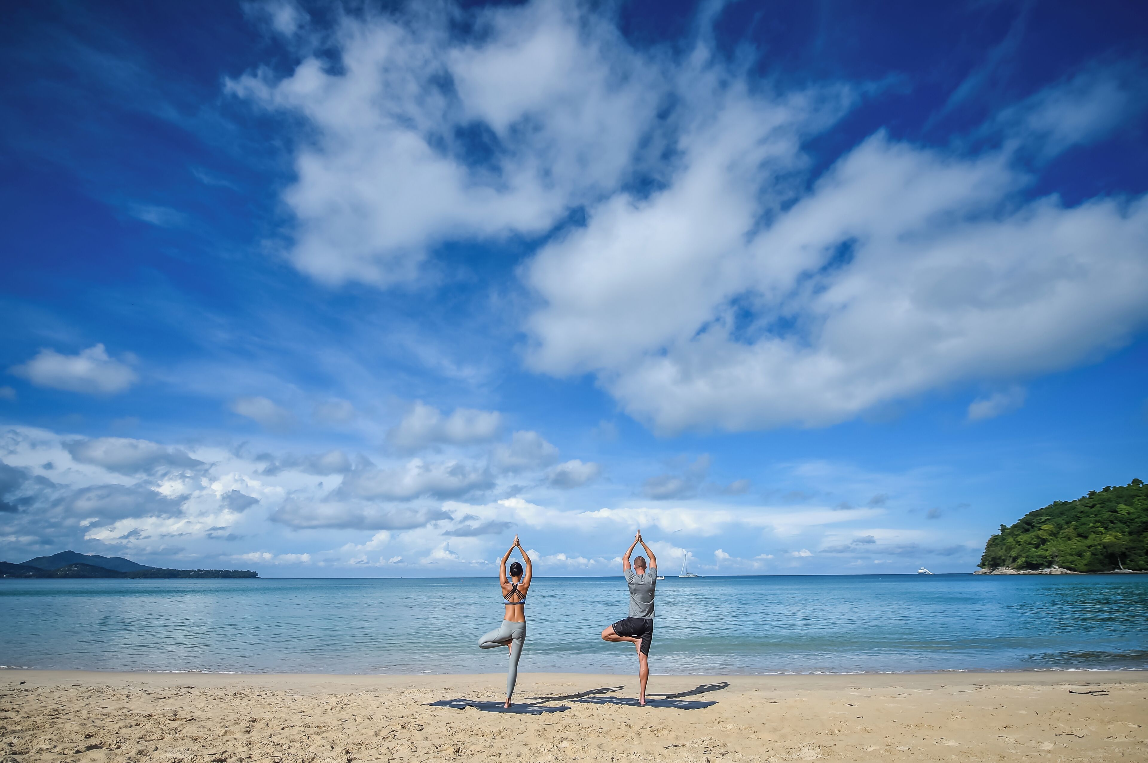 Two individuals performing yoga on a beach with a clear sky and calm sea in the background.