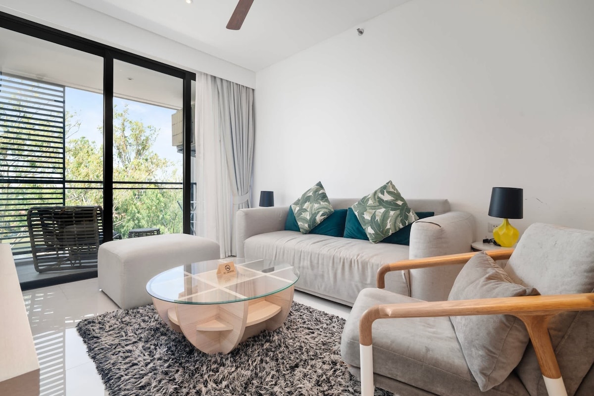 A modern living room with a glass coffee table, a sofa with patterned cushions, a wooden armchair, and a view through large windows.