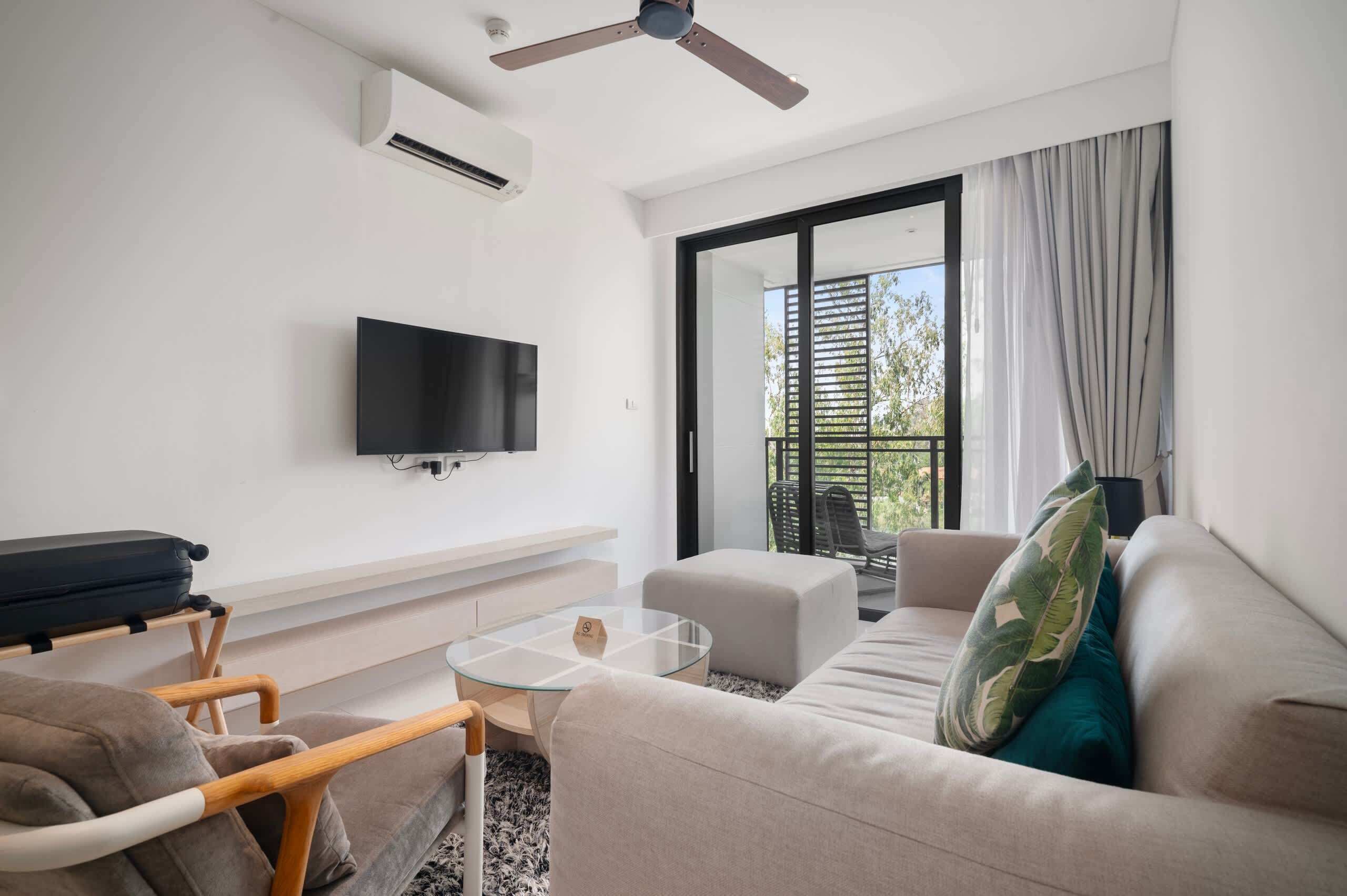 A modern living room with a flat-screen TV, a ceiling fan, a sofa, a glass coffee table, and a view of the outdoors through a sliding glass door.