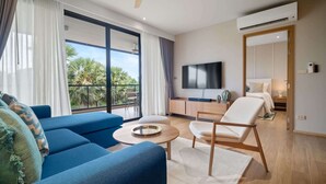 A modern living room with a sofa, armchair, and coffee table. A TV is mounted on the wall, and there's a balcony with a view of palm trees. - Laguna Beachside Residences Building 3 by Banyan Living (Choeng Thale)
