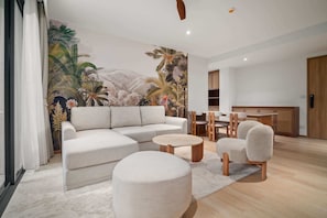 Una sala moderna con un sofá modular, una mesa de centro redonda, un reposapiés, una zona de comedor con sillas de madera y una cocina al fondo.