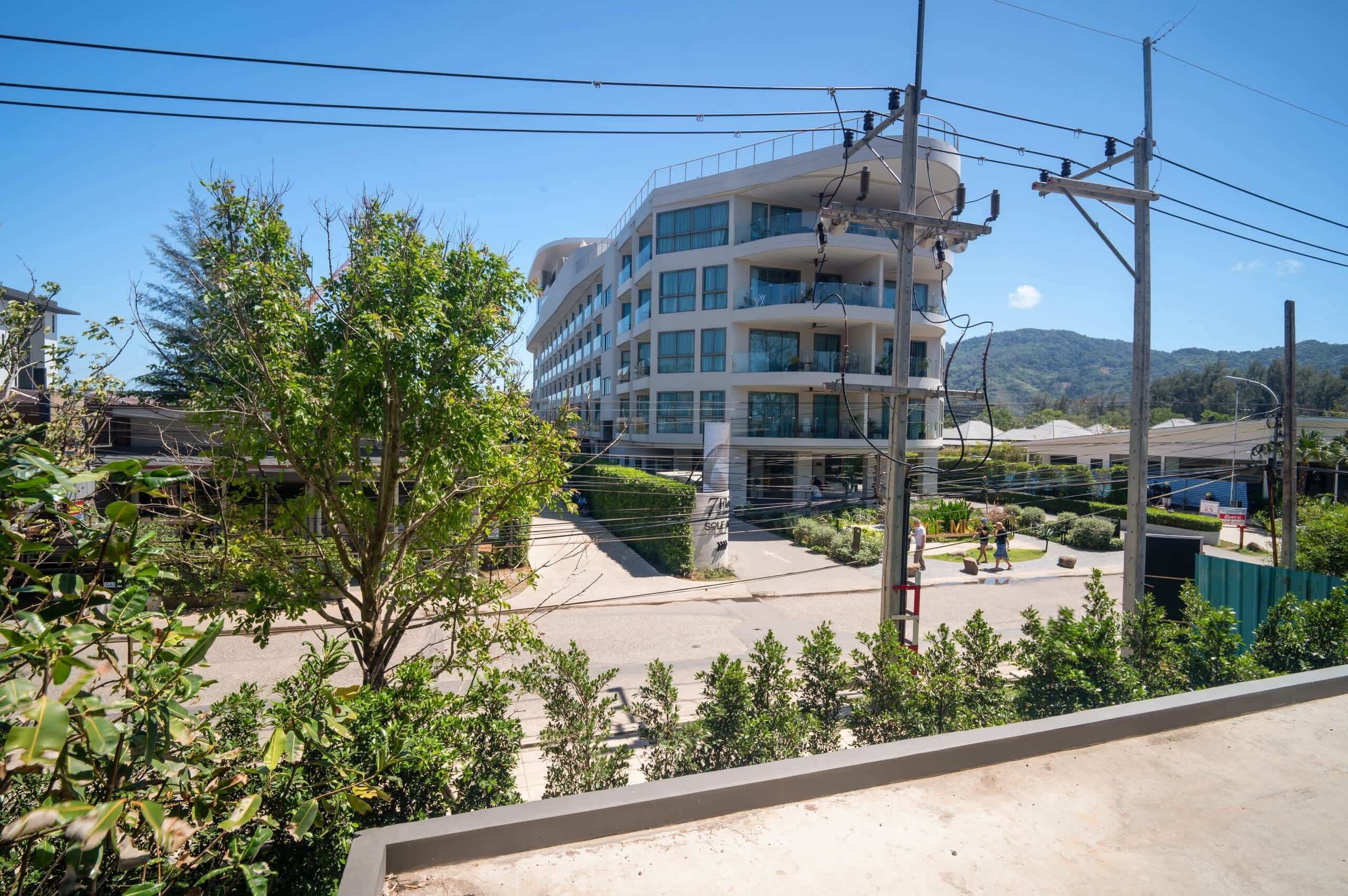 A multi-story building with balconies, surrounded by greenery and power lines.