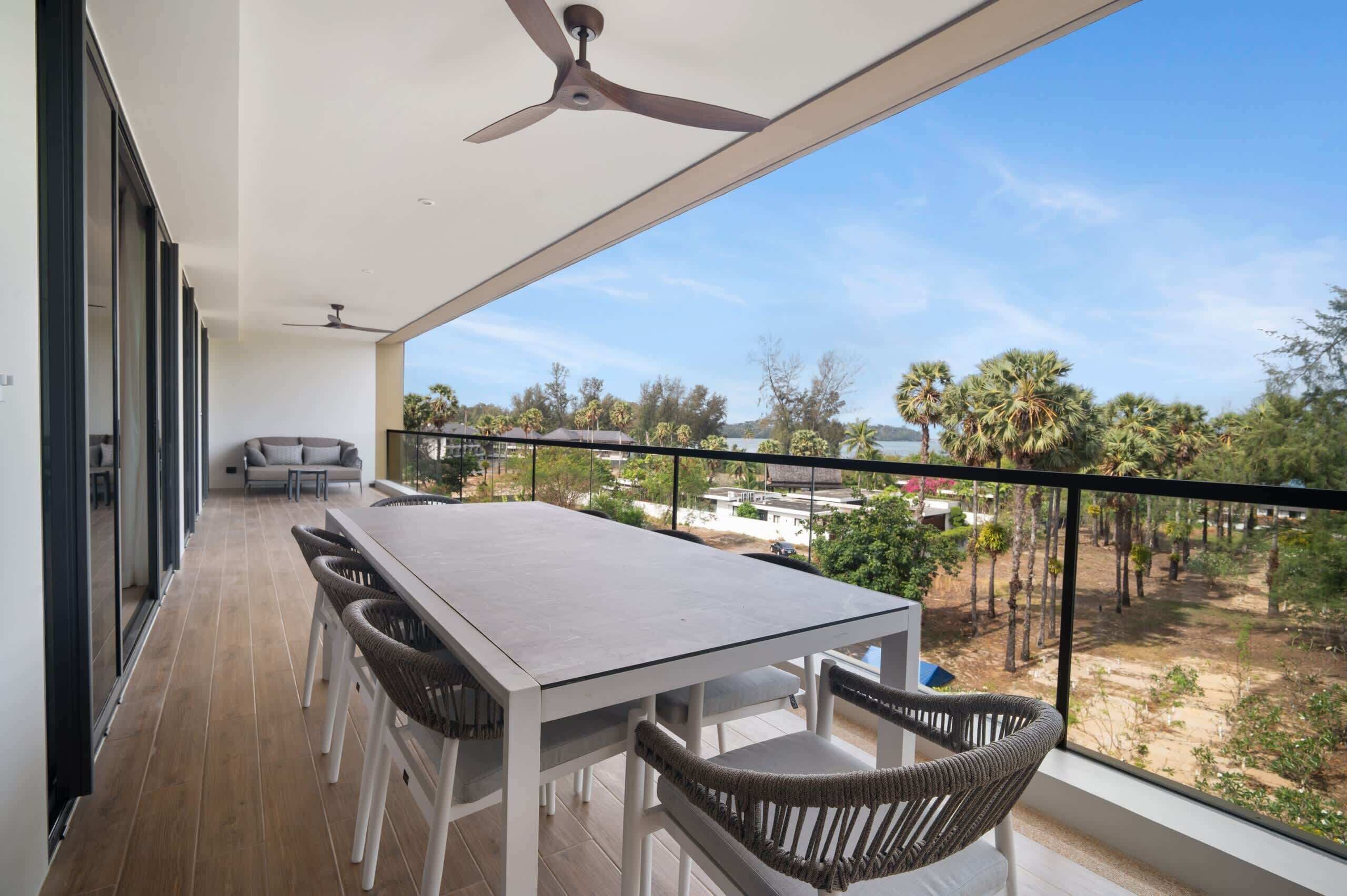 Een balkon met een eettafel en stoelen, een plafondventilator en uitzicht op palmbomen en gebouwen.