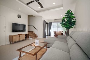 A modern living room with a sofa, a coffee table, a TV, a staircase, and a large plant. - Laguna Parkside Residences by Banyan Living (Choeng Thale)