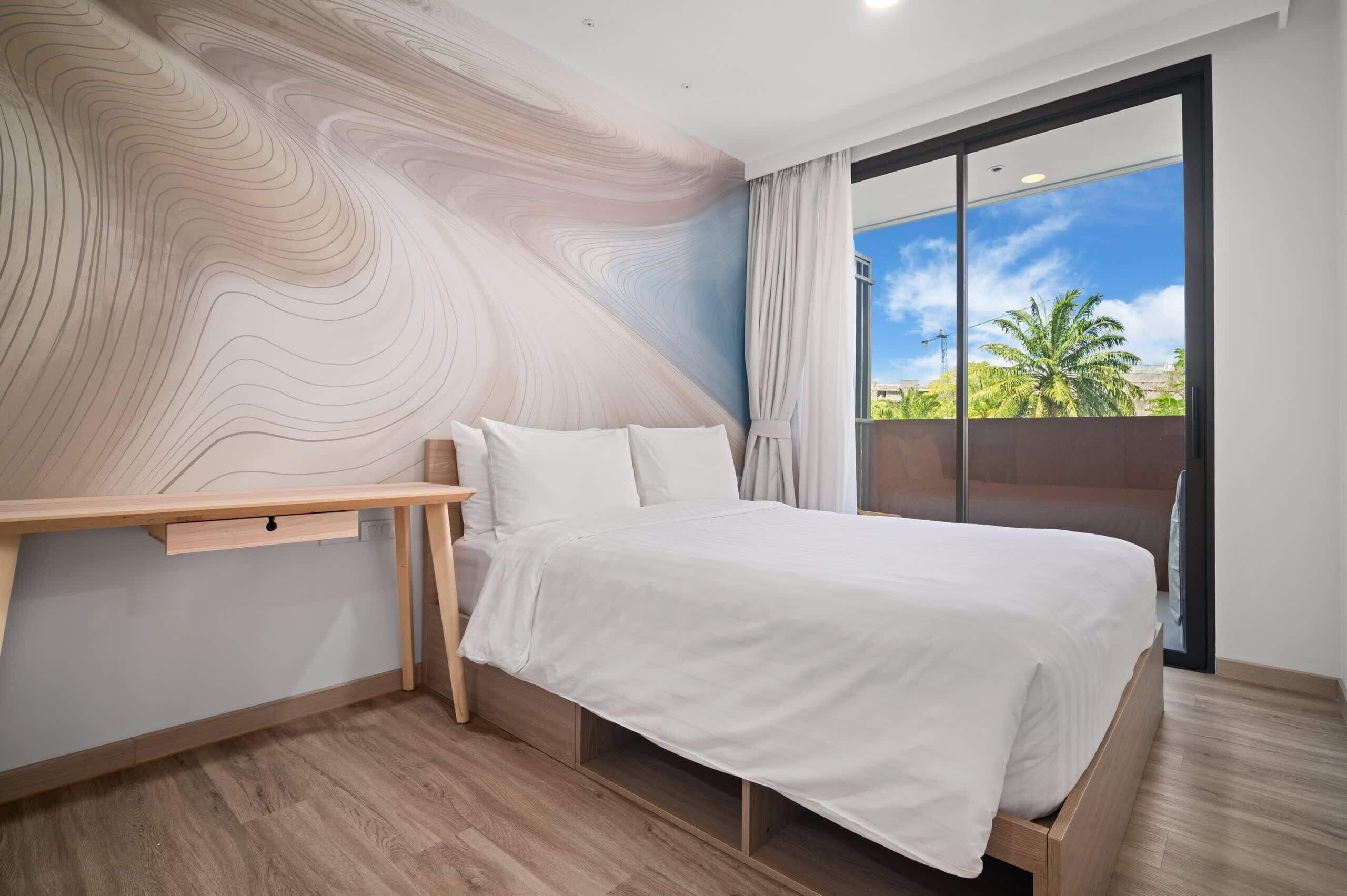Quarto moderno com escrivaninha de madeira, cama com roupa de cama branca e uma janela grande com vista para palmeiras.