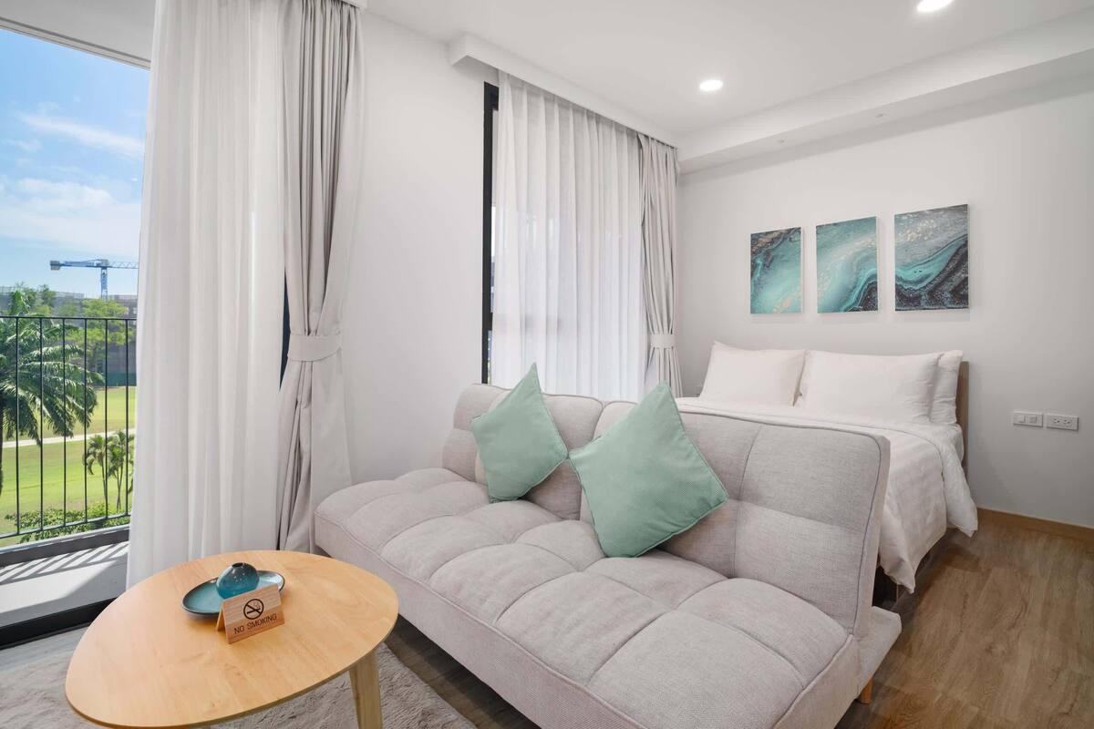 A modern hotel room with a sofa, a small table, a bed, and a balcony with a view of palm trees and a construction crane.