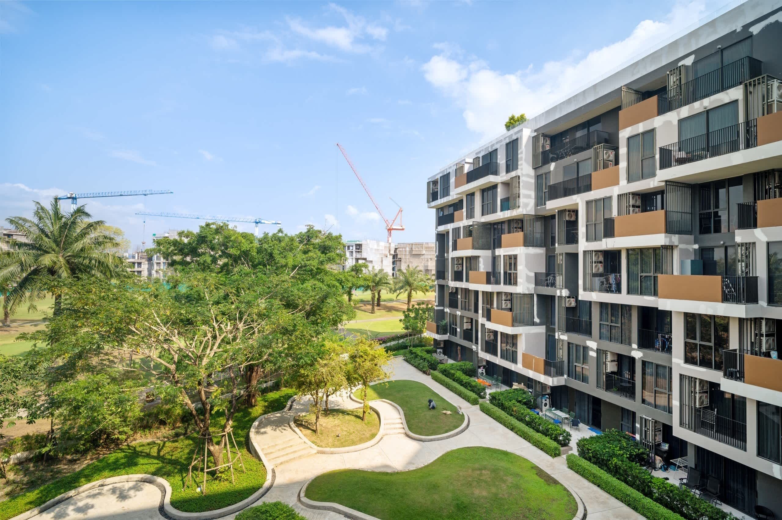 Un moderno edificio residencial de varios pisos con balcones, un jardín ajardinado y grúas de construcción al fondo.