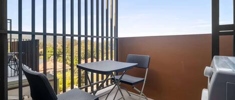 Een balkon met een tafel, twee stoelen, een barbecue en uitzicht op bomen en gebouwen.