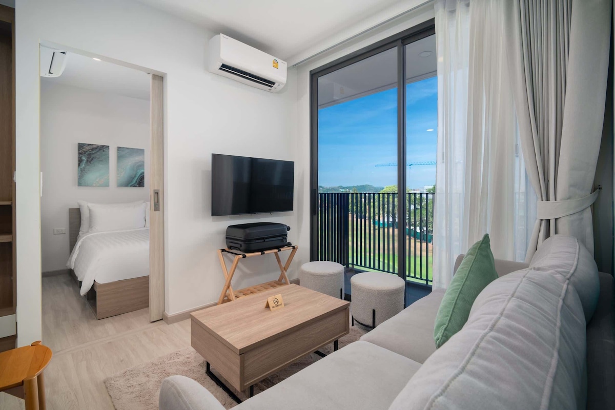 A modern hotel room with a bed, a flat-screen TV, a wooden coffee table, a sofa, and a balcony with a view.