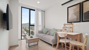 Un salon moderne comprenant un canapé, une table basse, une table à manger avec des chaises, un téléviseur fixé au mur et de grandes fenêtres offrant une vue sur les bâtiments et les collines.
