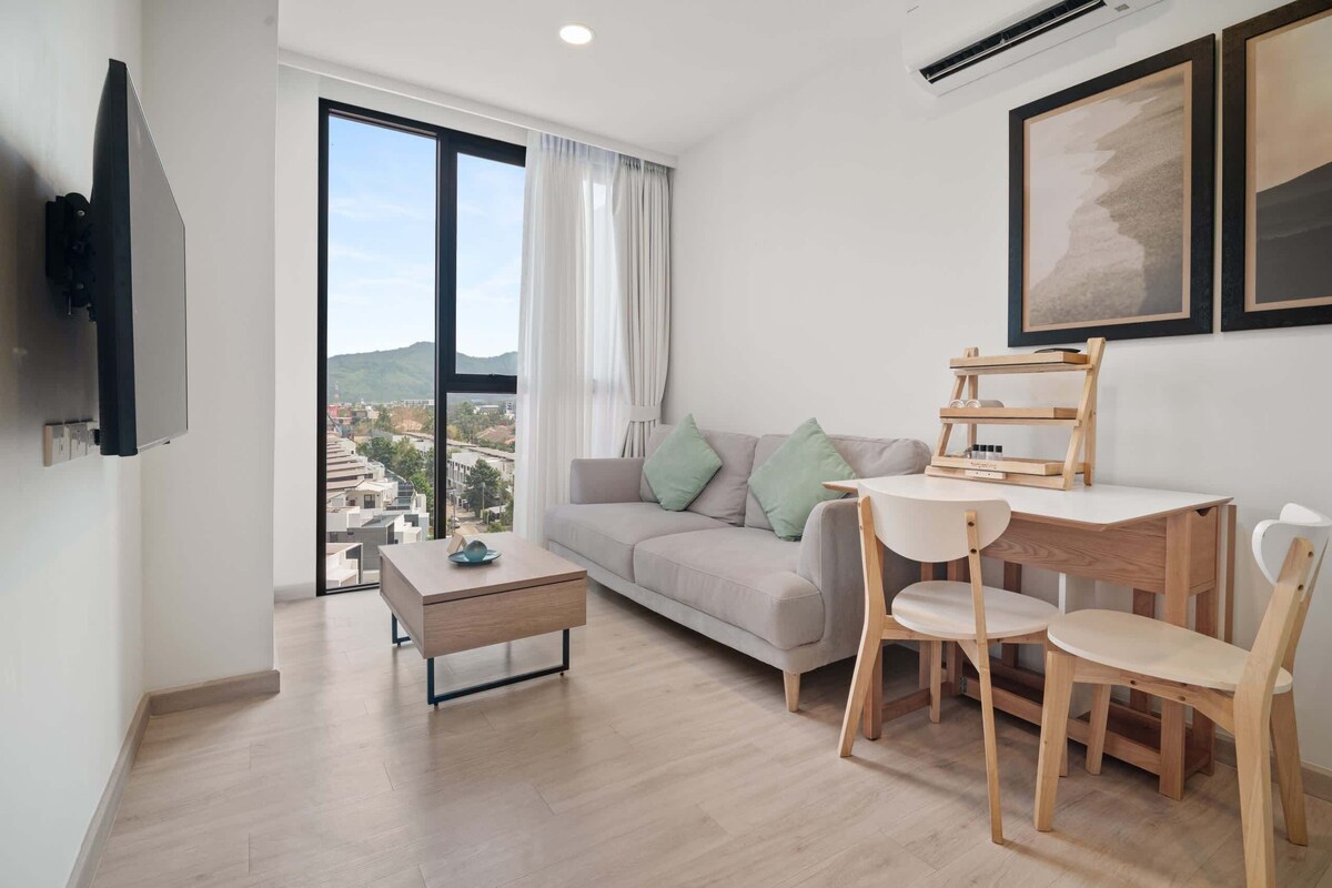 A modern living room with a sofa, a coffee table, a dining table with chairs, a TV mounted on the wall, and large windows with a view of buildings and hills.
