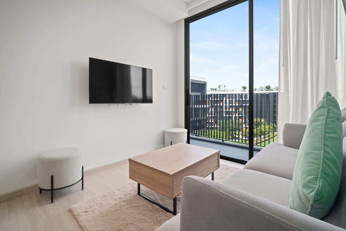 A modern living room with a flat-screen TV, a light-colored sofa, a wooden coffee table, and a small round stool.