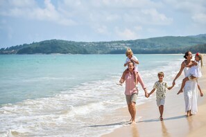 Eine Familie spaziert am sandigen Strand mit freiem Blick auf das Meer und hügelige Landschaften im Hintergrund.