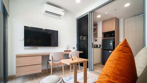 Ett modernt vardagsrum med en platt-TV, ett skrivbord med stolar, ett litet bord och en soffa med en orange kudde.