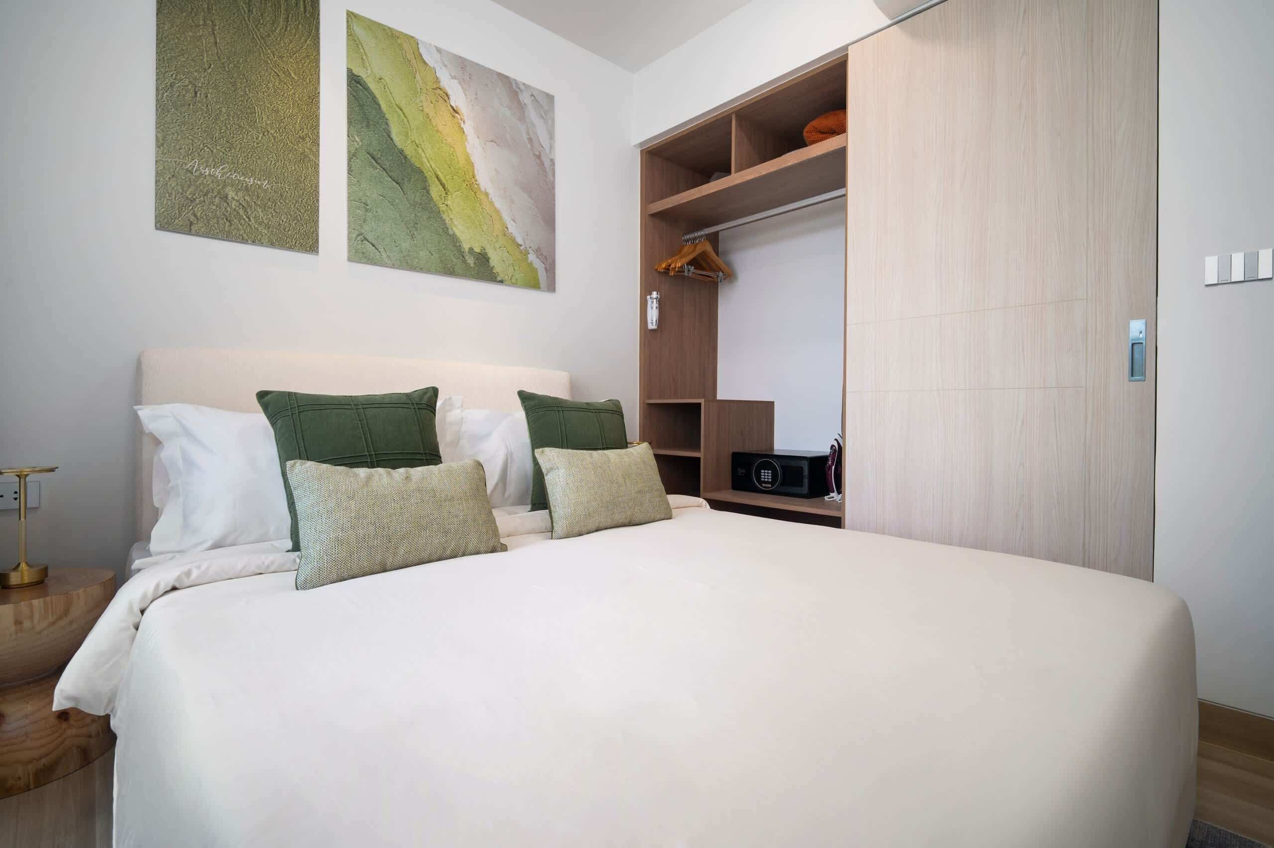 Un dormitorio con cama, almohadas, una mesita de noche, un armario y una obra de arte colgada en la pared.