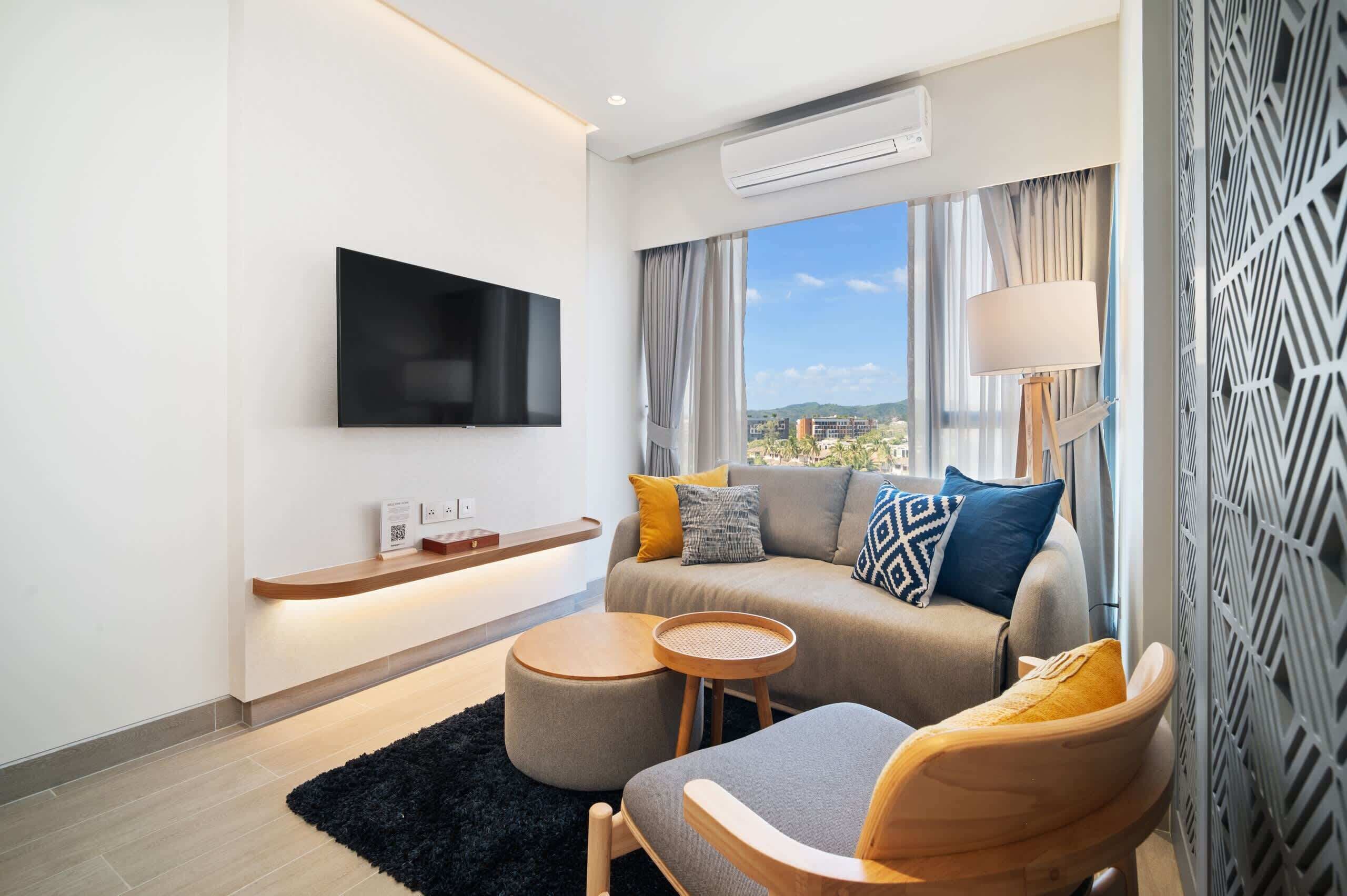 Een moderne woonkamer met een flatscreen televisie, een bank met kussens, een ronde houten tafel en stadszicht door grote ramen.