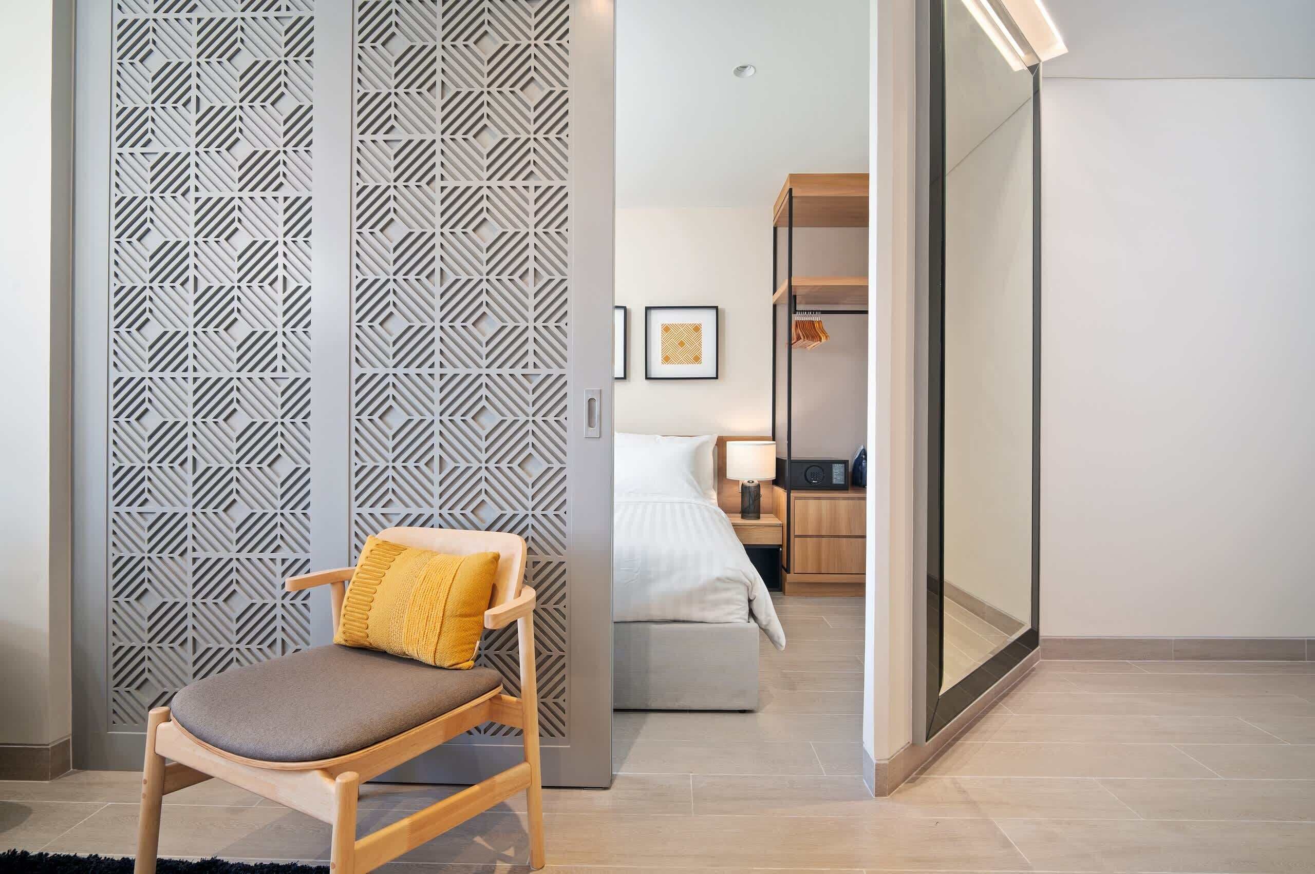 A hotel room with a bed, bedside table, lamp, and a chair with a cushion.