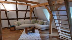 A loft-style room with exposed wooden beams, a white sofa with green cushions, a small wooden table, and a ladder. - Maison des Vergers, nature meets craftsmanship (Hoffen)