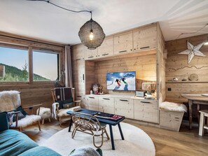 Ein gemütliches Blockhäuschen mit Holzwänden, einem großen Fenster, einem Fernseher, einer Essecke und einer Sternen-Dekoration.