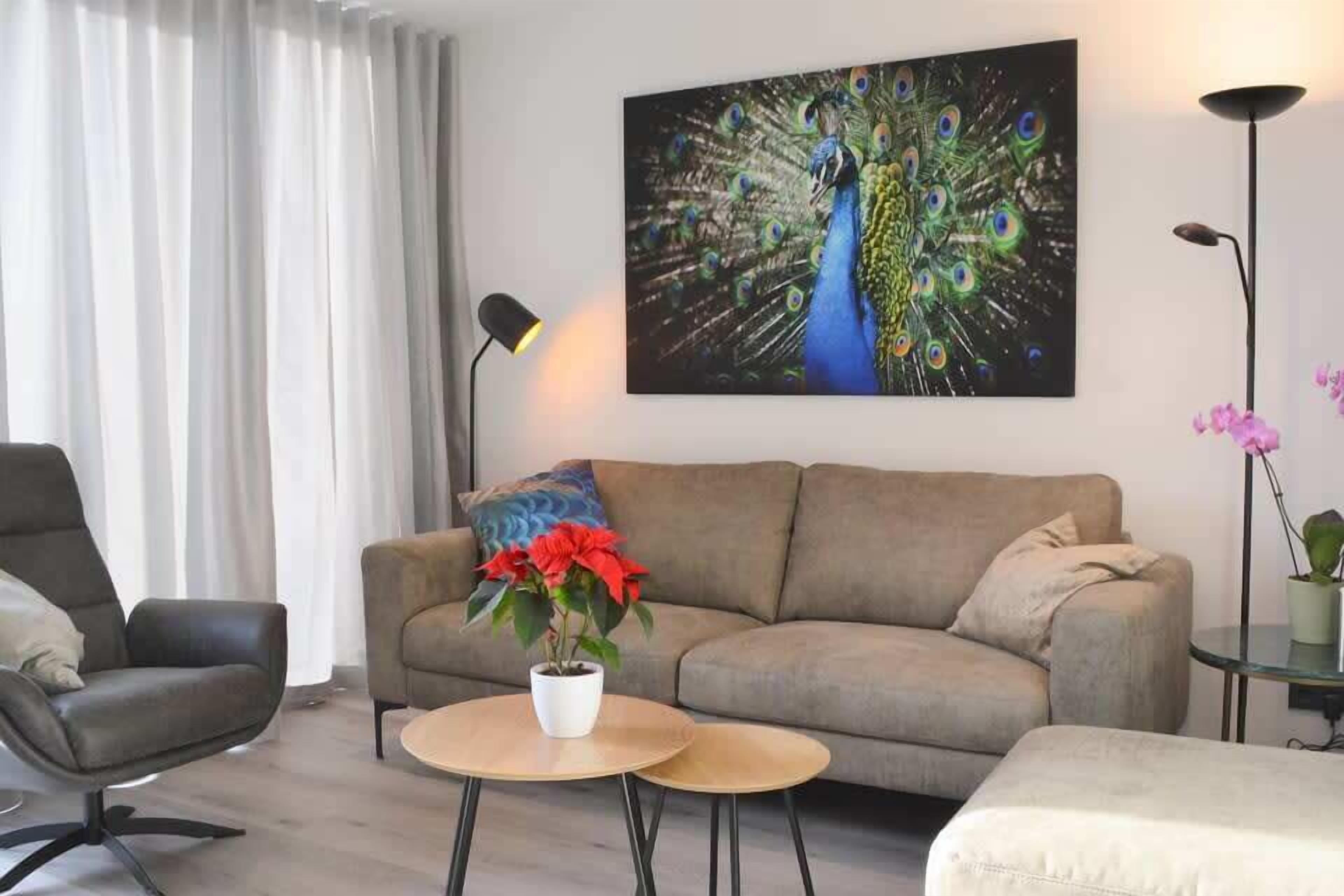 Ein Wohnzimmer mit einer Couch, einem Sessel, einem runden Tisch mit einem Blumentopf und einem Pfau-Gemälde an der Wand.