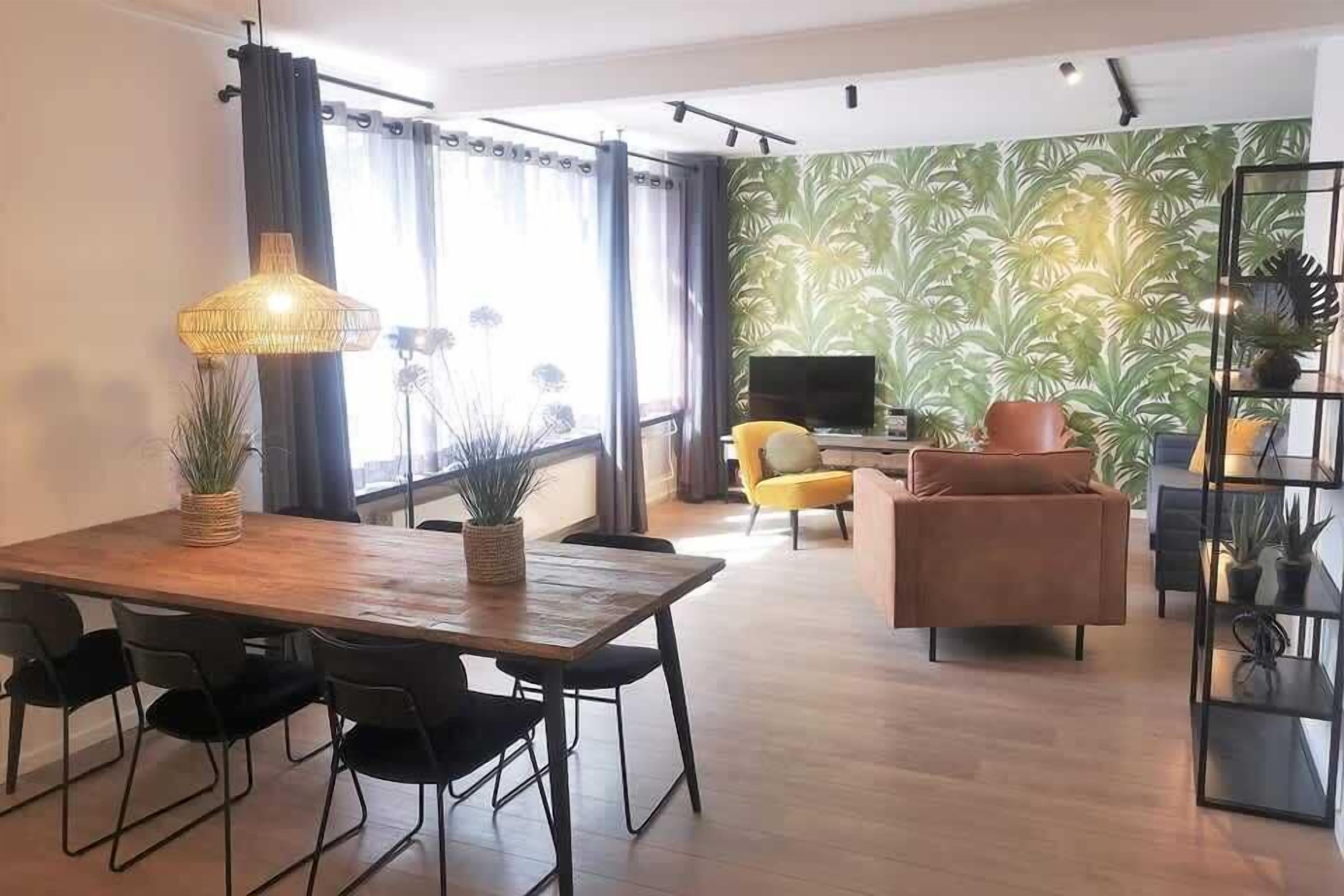 Ein modernes Wohnzimmer mit einem Holzesstisch, schwarzen Stühlen, einem gelben Sessel, einem braunen Sofa, einem Fernseher und einer Wand mit grünem Muster.