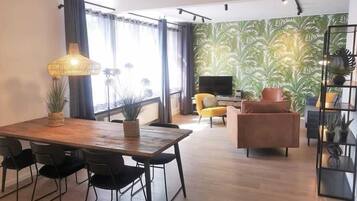 Ein modernes Wohnzimmer mit einem Holzesstisch, schwarzen Stühlen, einem gelben Sessel, einem braunen Sofa, einem Fernseher und einer Wand mit grünem Muster.