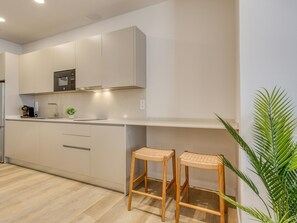 Eine moderne Küche mit weißen Schränken, einem eingebauten Backofen und einer Kaffeemaschine. Zwei hölzerne Barhocker mit gewobenen Sitzflächen.