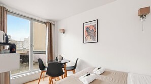 Ein Hotelzimmer mit Bett, einem Esstisch mit Stühlen, einem Fenster mit Vorhängen, einem gerahmten Bild an der Wand und einer Kaffeemaschine auf der Anrichte.