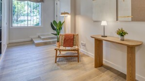Una habitació àmplia amb terra de fusta, una cadira amb una manta, una làmpada de peu, una consola amb una planta i un gran finestral amb vista a vegetació.