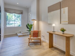 Ein geräumiger Raum mit Holzboden, einem Sessel mit Decke, einer Stehlampe, einem Beistelltisch mit Pflanze und einem großen Fenster mit Blick auf Grünflächen.