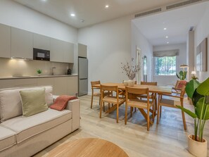 Ein modernes Wohnzimmer mit einer Couch, einem Esstisch mit Stühlen, einer Küchenzeile mit Kühlschrank und einem großen Fenster.
