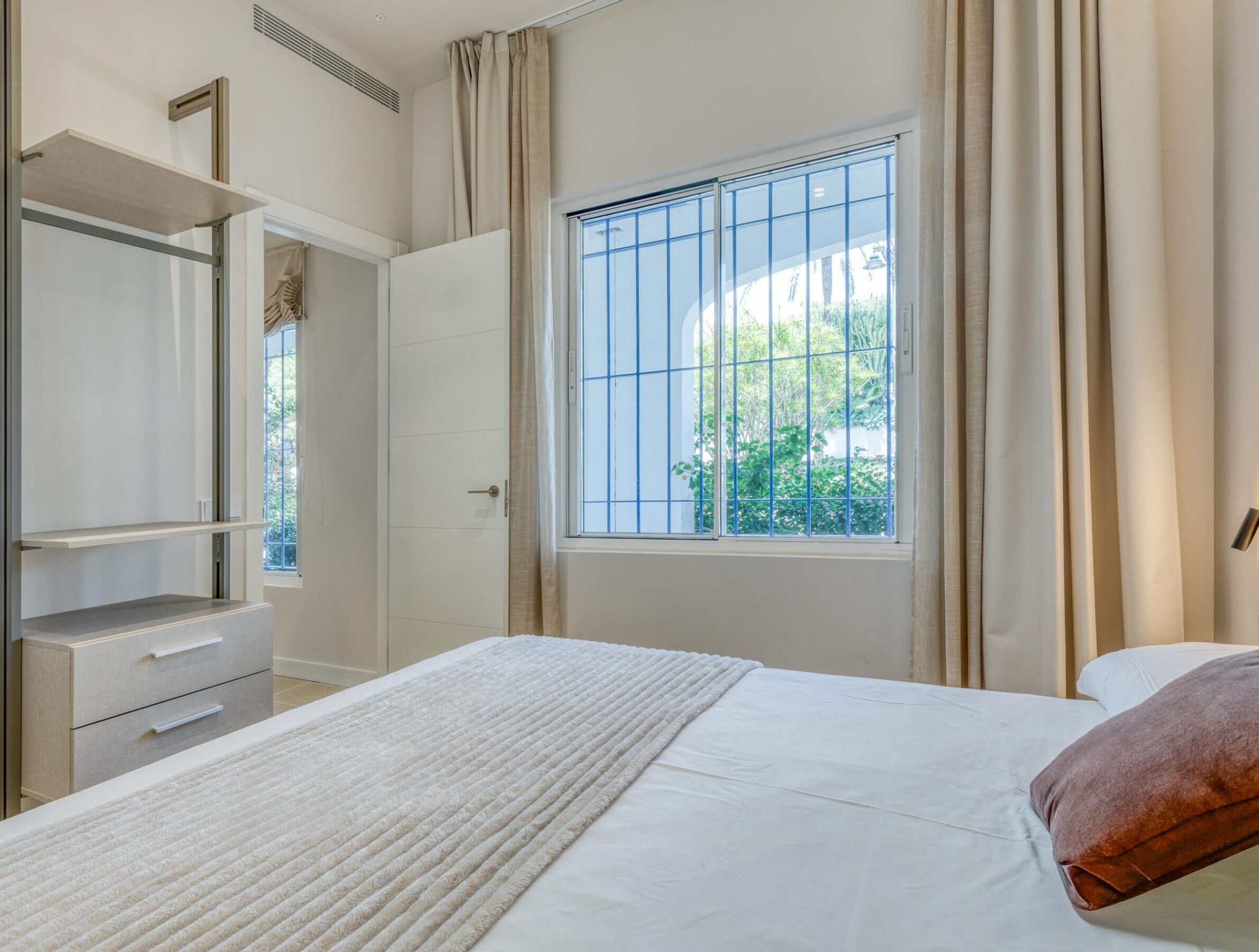 Une chambre avec un lit, une armoire, une fenêtre avec des barreaux et une vue sur la verdure à l’extérieur.