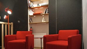 Une pièce comprenant deux fauteuils rouges, une petite table ronde, une bibliothèque, un extincteur et une chaise en bois.