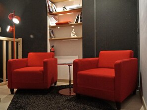 Une pièce comprenant deux fauteuils rouges, une petite table ronde, une bibliothèque, un extincteur et une chaise en bois.