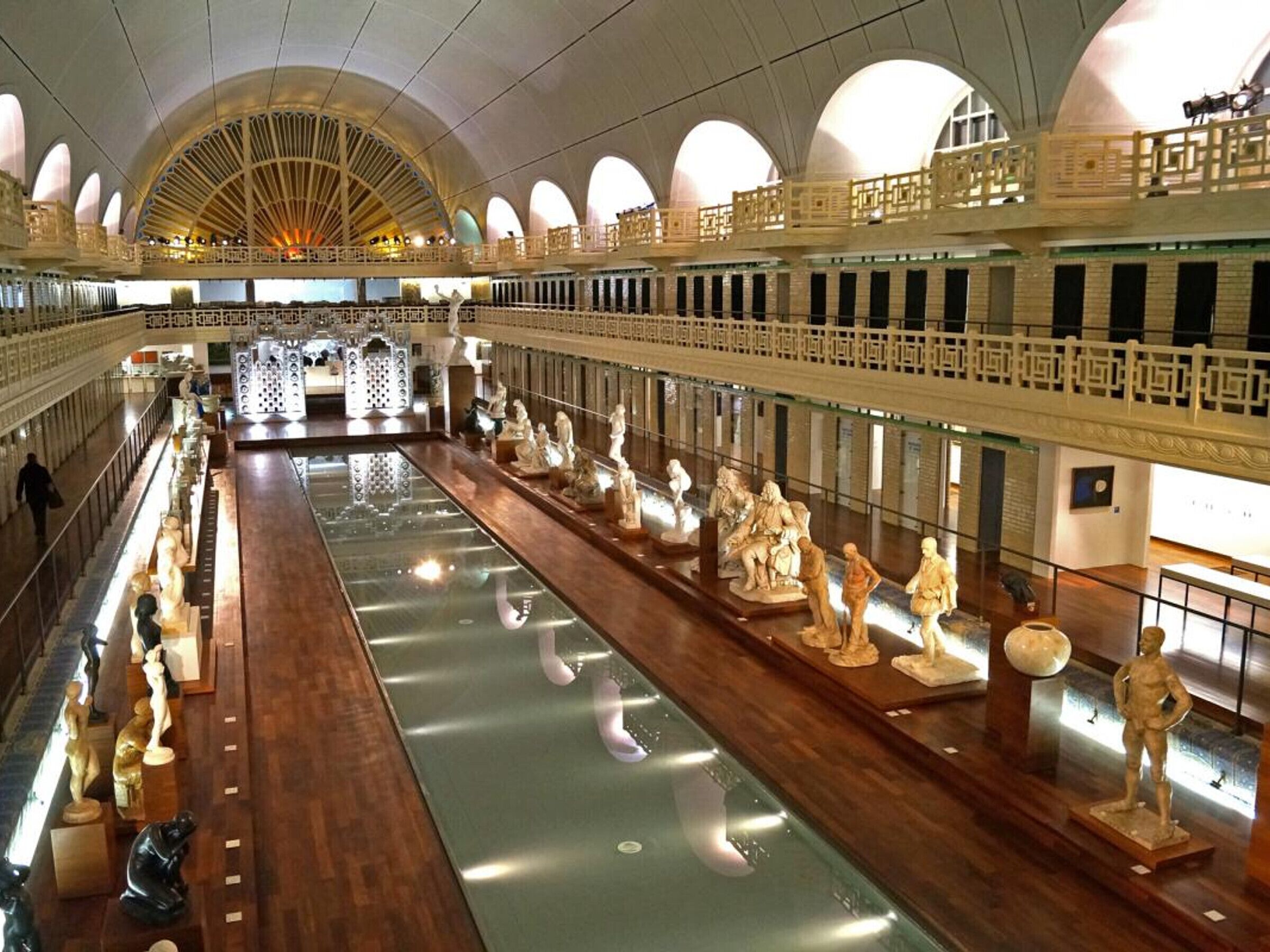Un interior espacioso con una fuente de agua central reflectante, varios niveles con balcones y esculturas clásicas en exhibición.